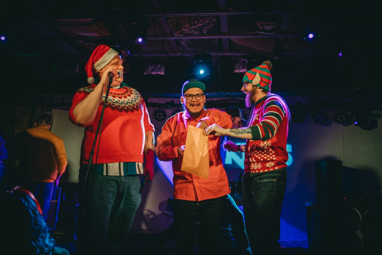 Performers in colorful winter sweaters singing together on stage during a holiday-themed concert.