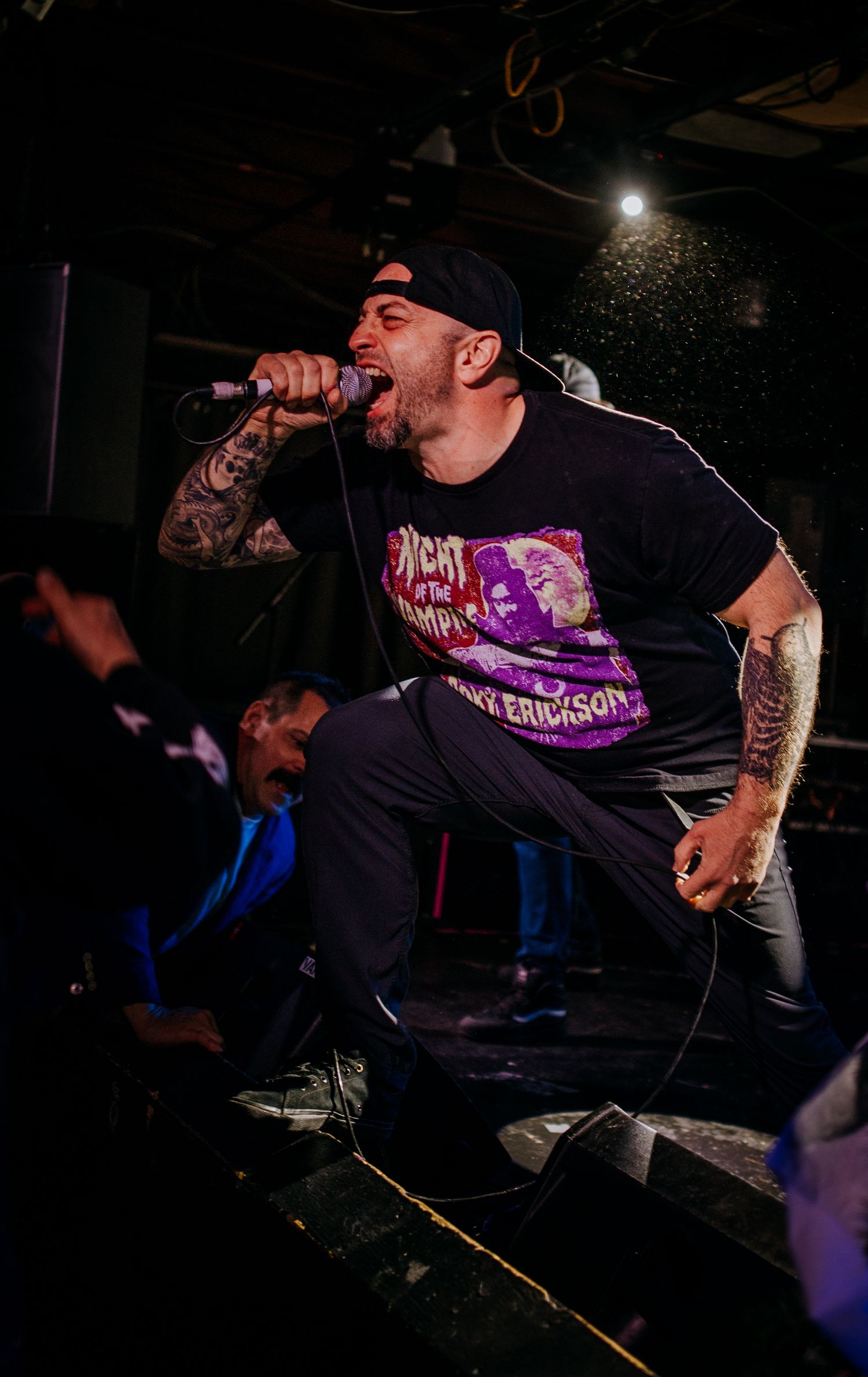 Intense stage performance with dramatic lighting showing a vocalist in a black graphic t-shirt.