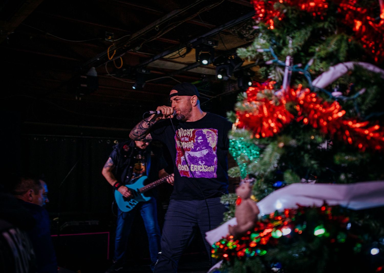 Musical performance with Christmas tree decorations visible and blue stage lighting.