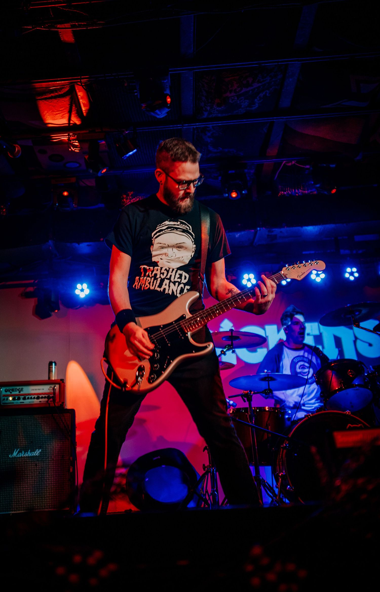 Guitar player performing on stage with red and blue lighting effects creating dramatic atmosphere.
