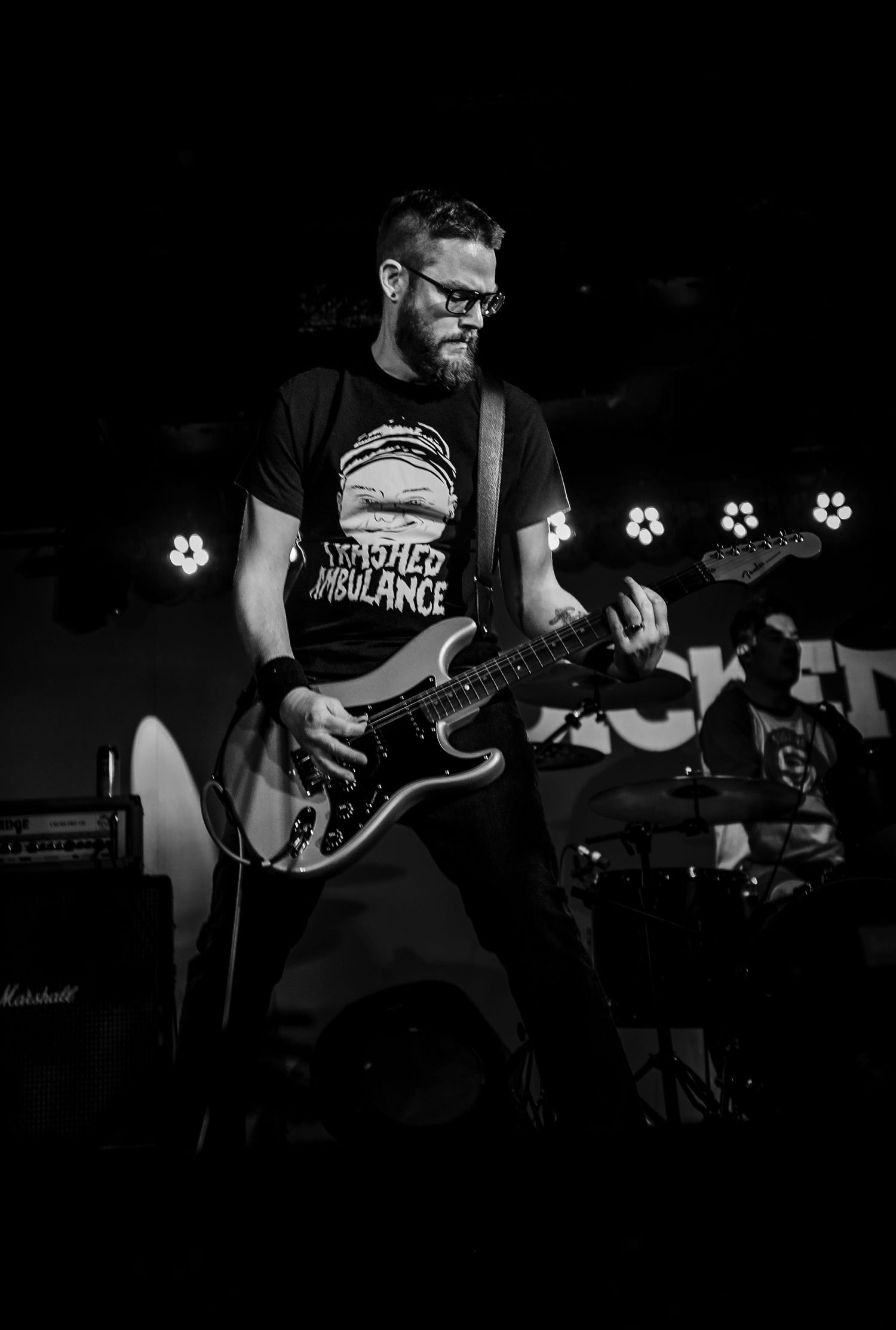 Black and white photo of guitarist in band t-shirt performing on dark stage with stage lights.