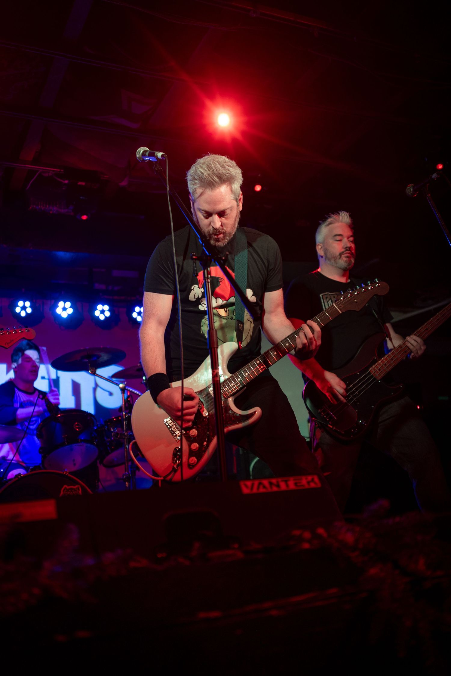 Guitarist performing energetically on stage with red stage lighting and band members in background.