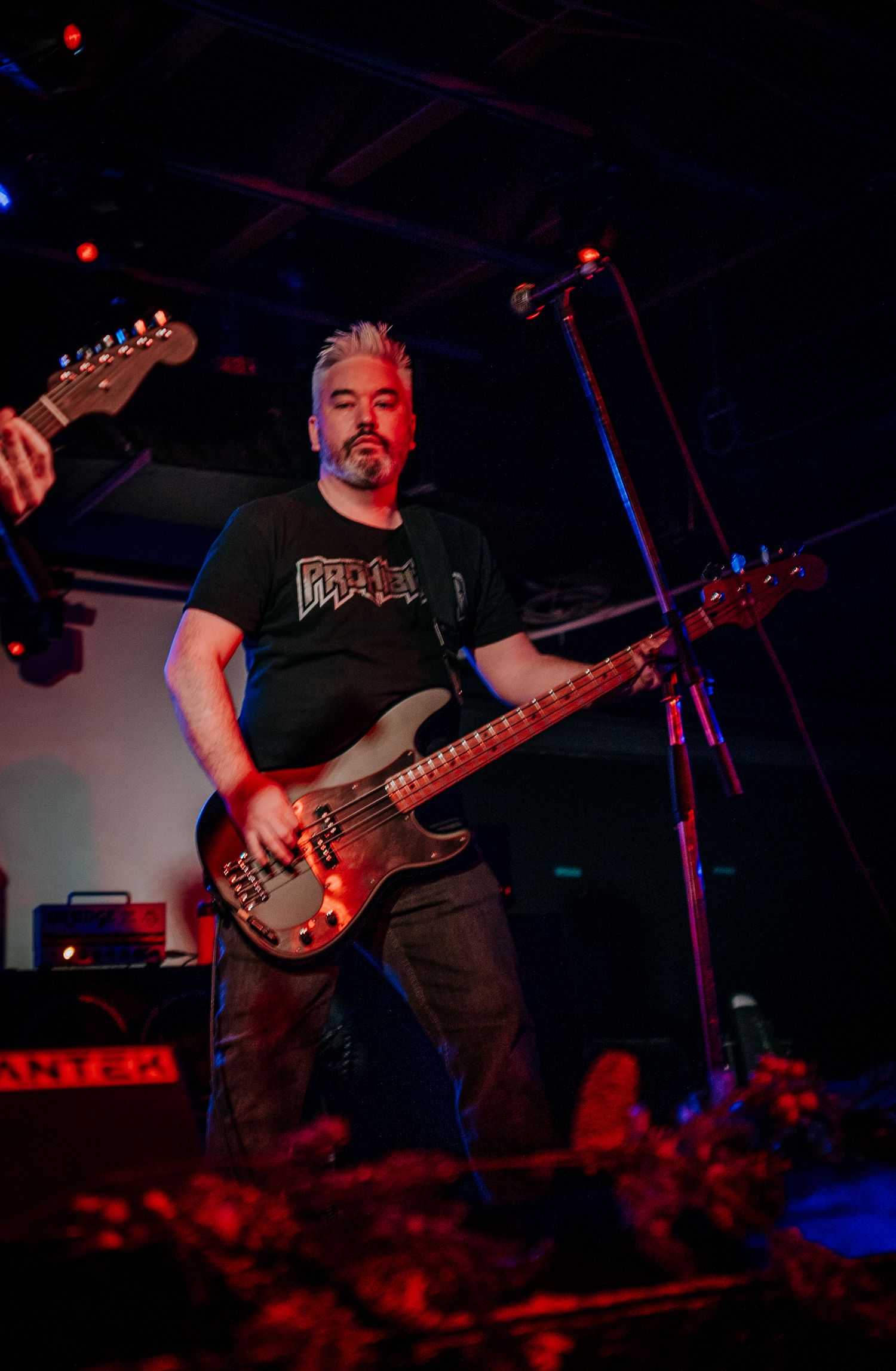 Bass guitarist in black band shirt performing under dramatic red stage lighting.
