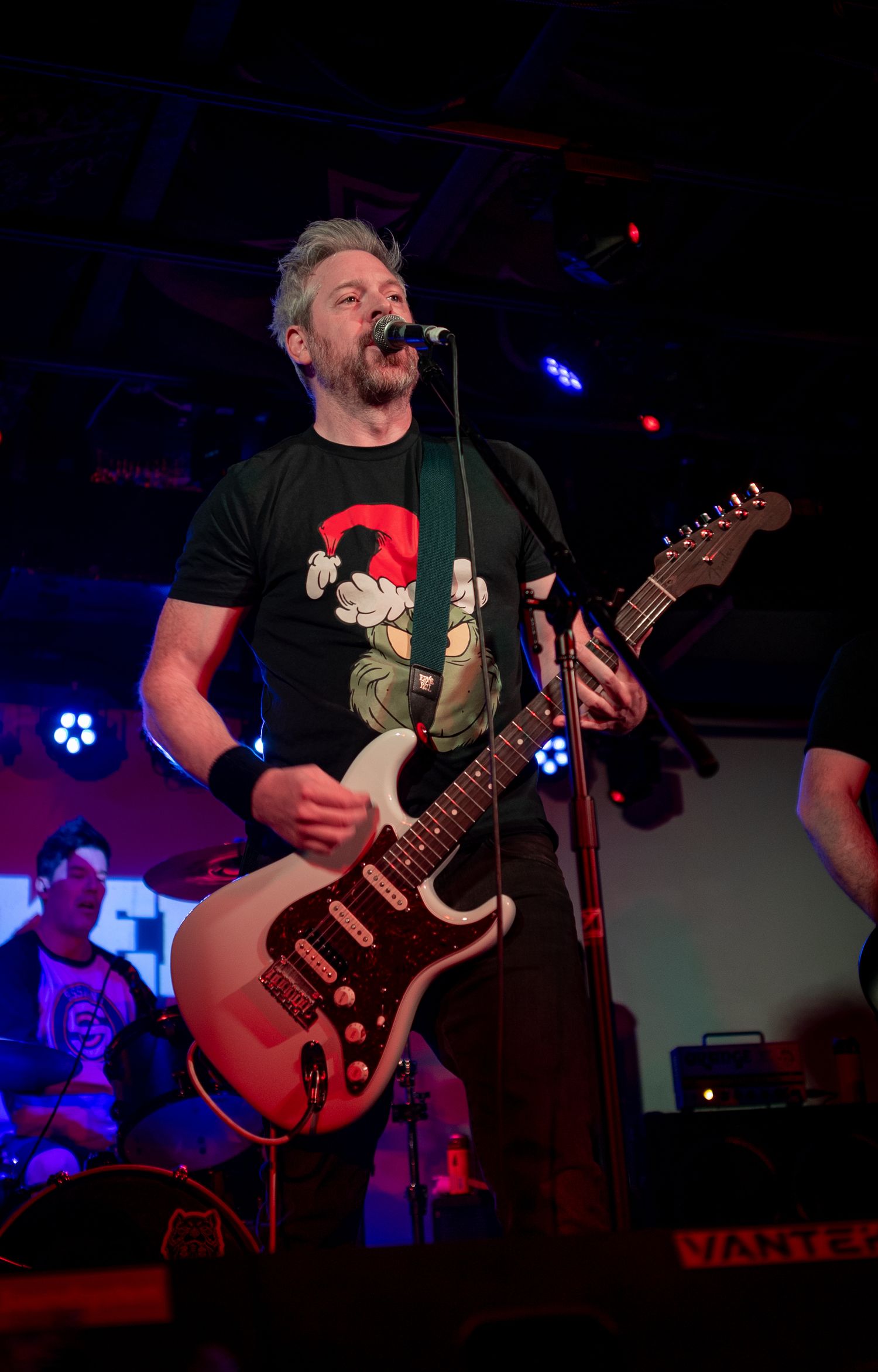 Musician playing red electric guitar wearing graphic t-shirt under blue stage lights.