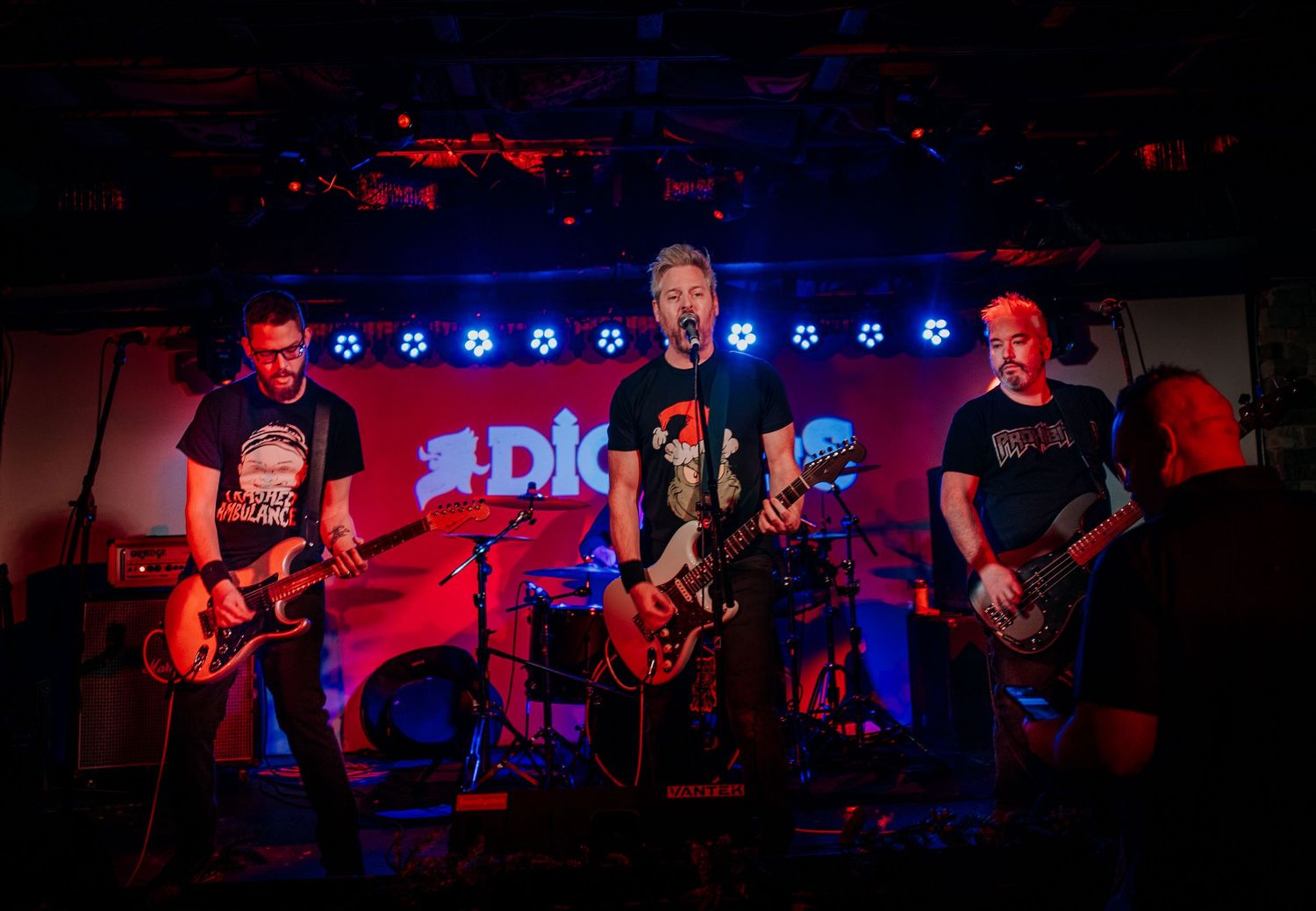 Rock band performing on stage with red and blue lighting effects and band logo visible.