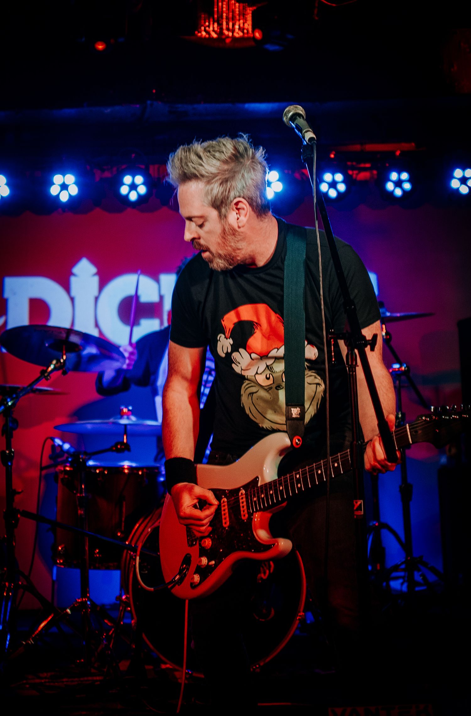 Musician playing red guitar on stage with blue LED lights and band logo in background.