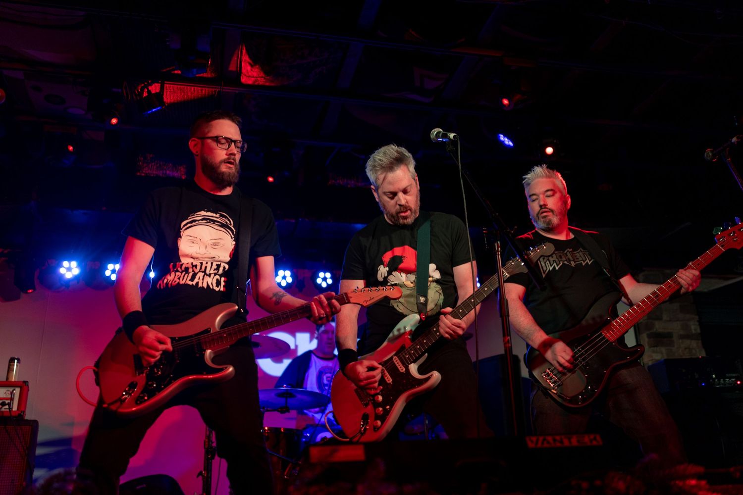 Band members playing electric guitars perform together on a dark stage with blue lighting effects.