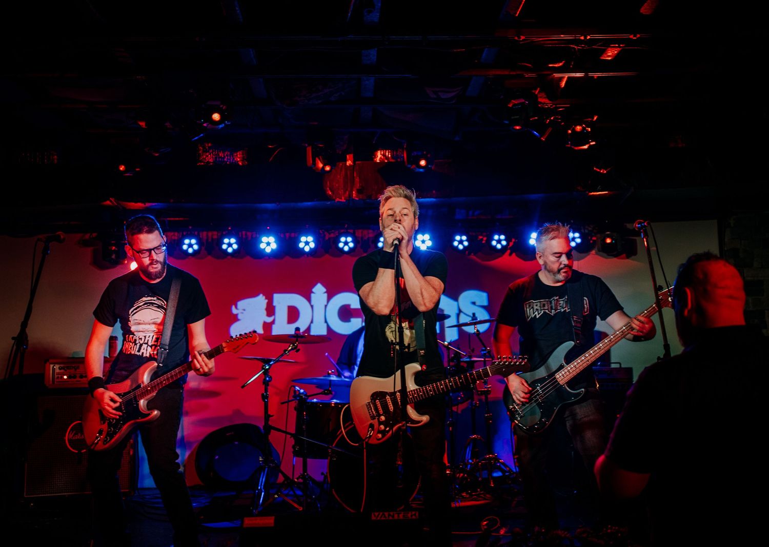 Rock band performs energetically on stage with illuminated signage and blue lighting in the background.