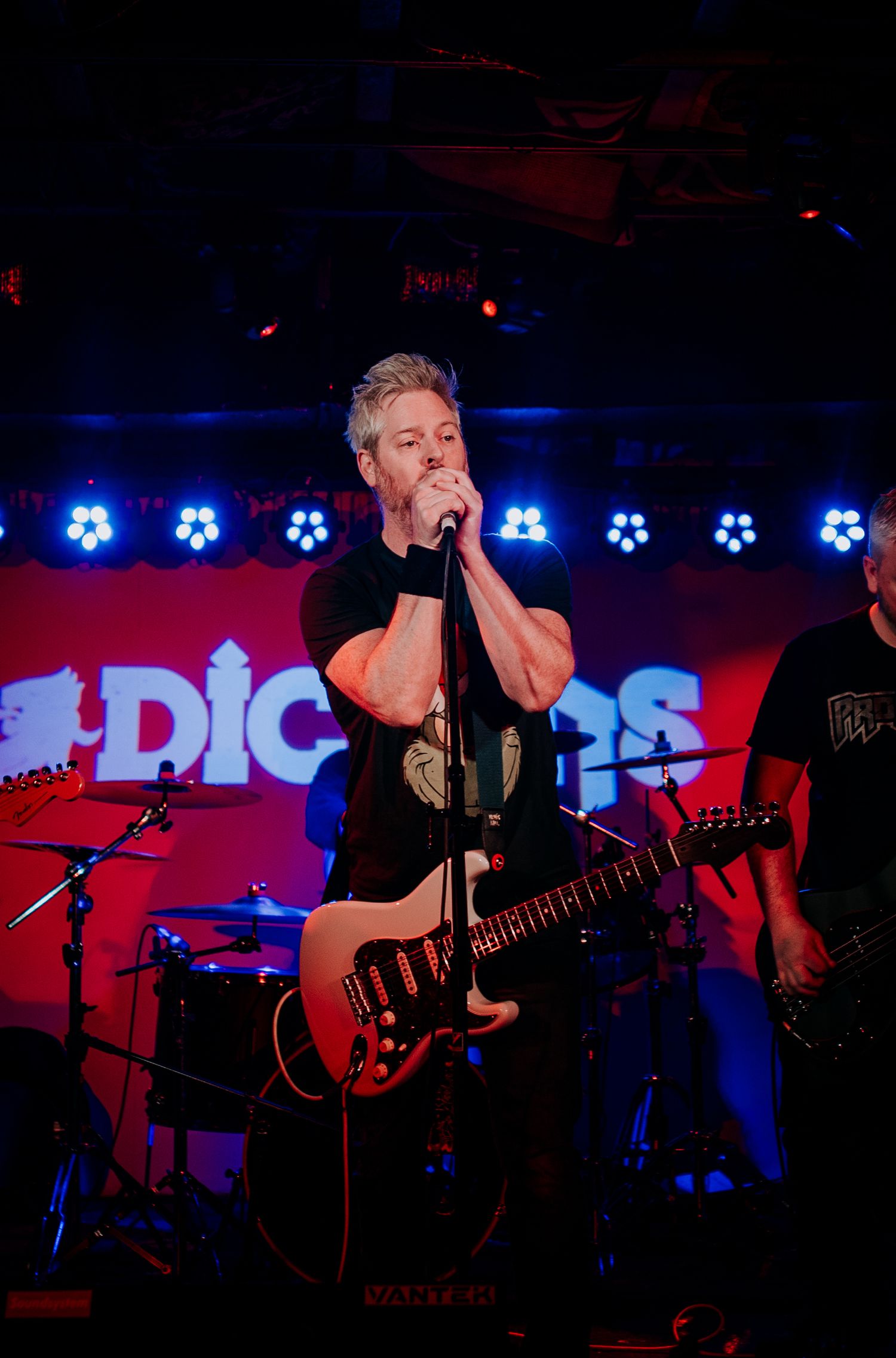 Musician sings into microphone while playing red electric guitar on dimly lit stage.