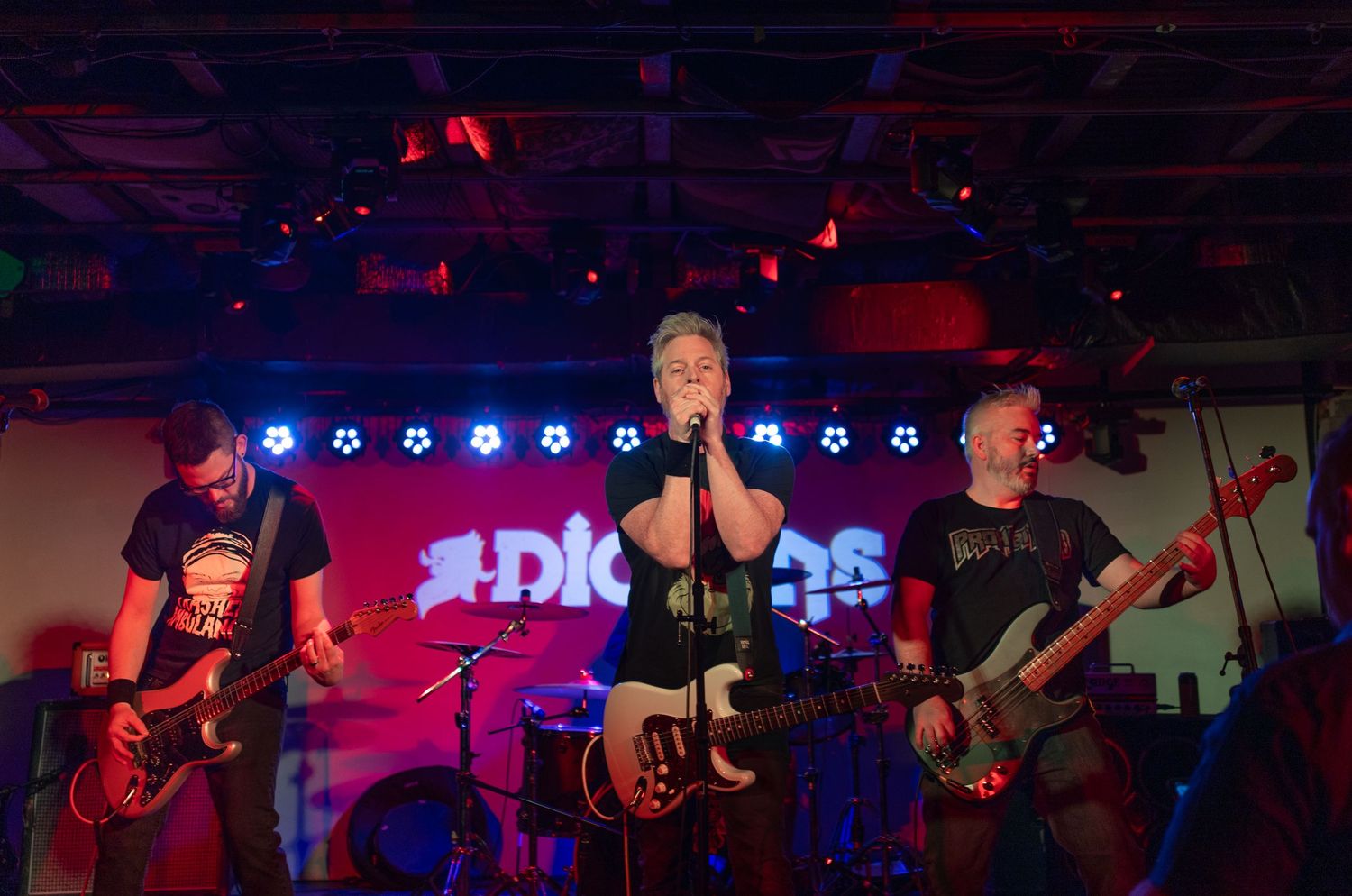 Rock band plays instruments on stage with blue accent lights and red ambient lighting.