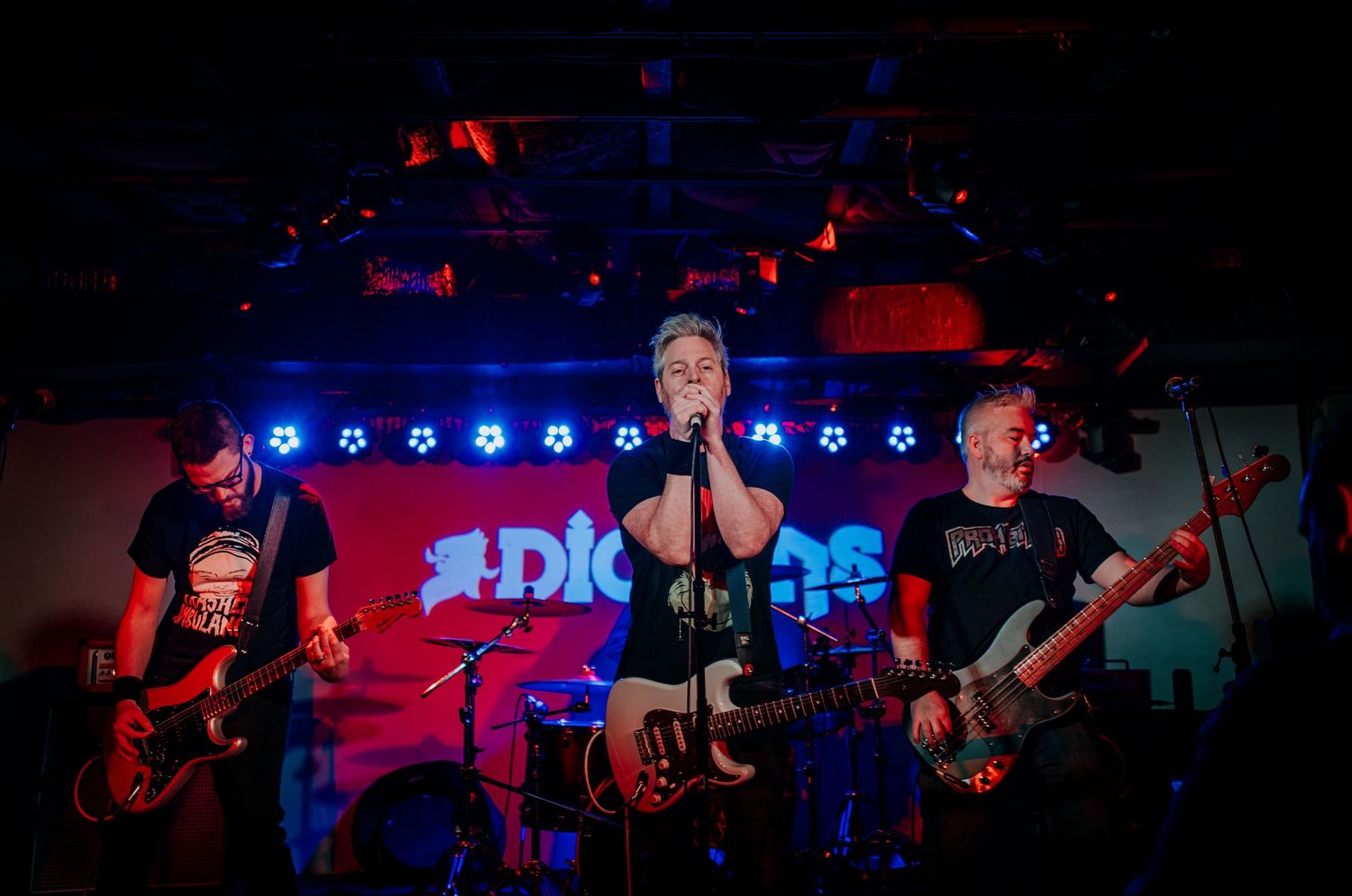 Three piece rock band performs on stage with dramatic lighting and venue signage visible.