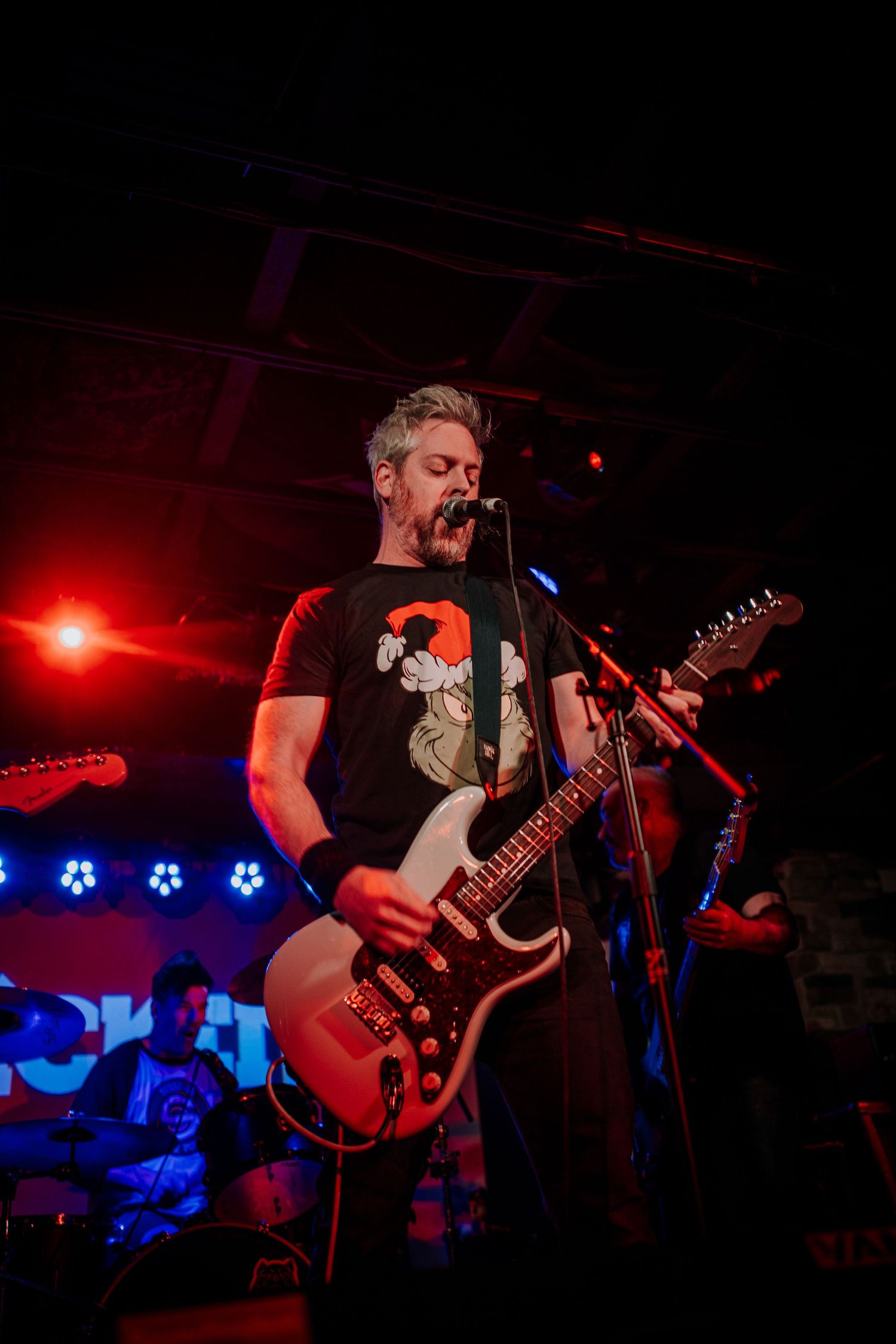 Musician plays red electric guitar while singing on dark stage with blue lighting effects.
