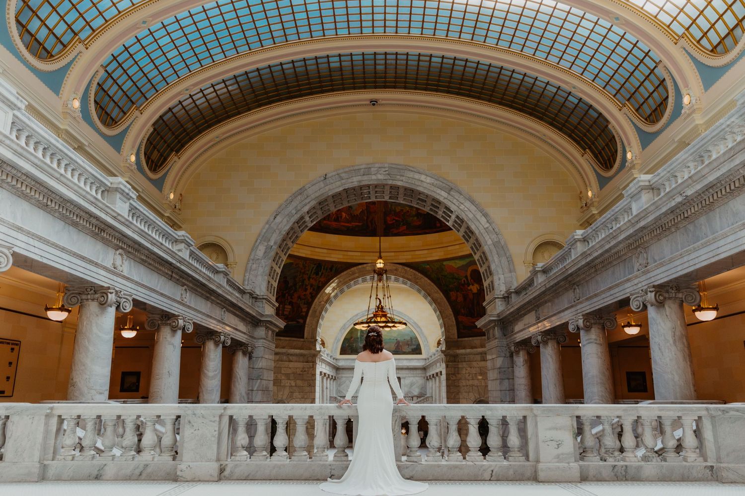 A bride poses in an elegant white dress inside a grand historic building with ornate architecture.