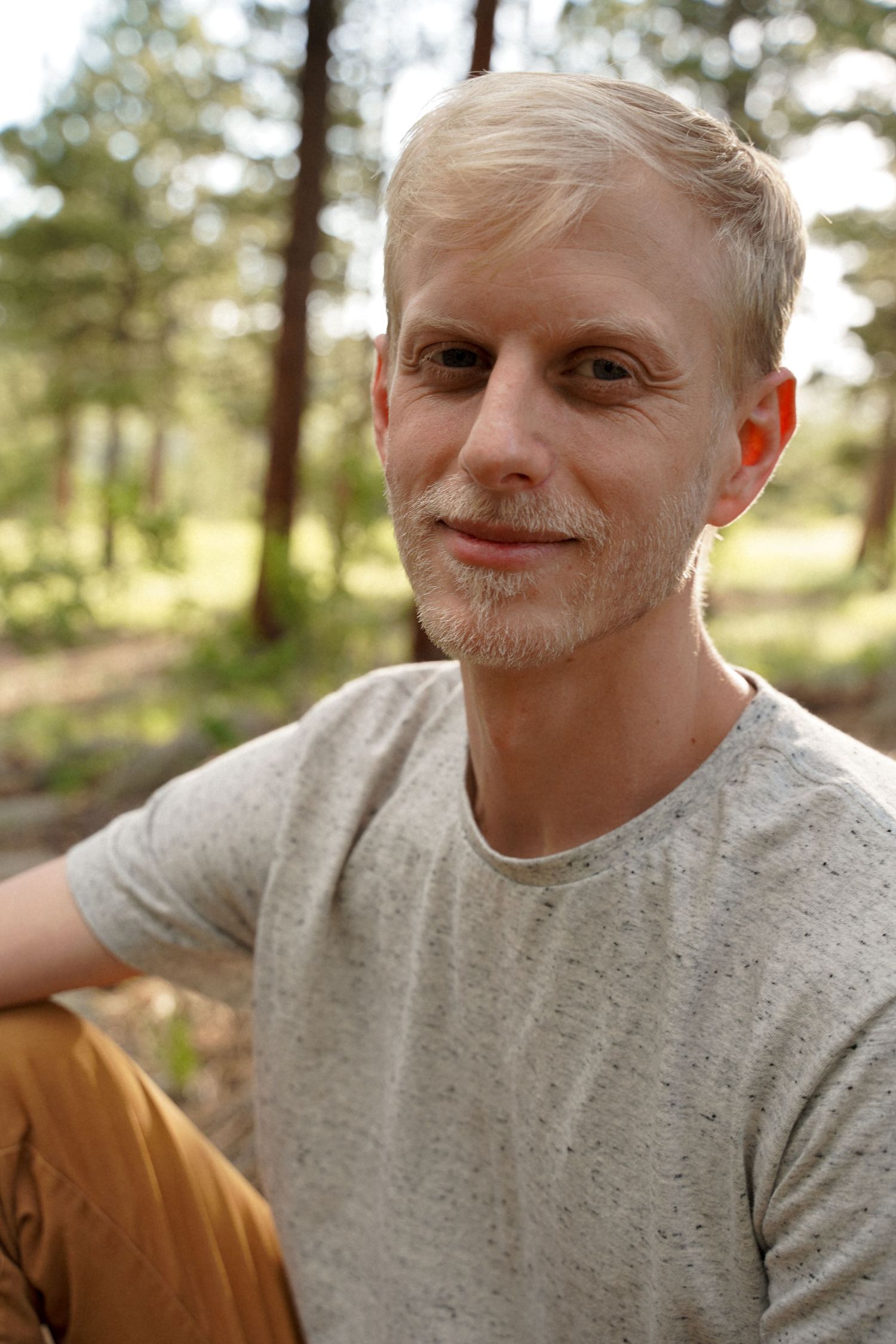 Natural outdoor portrait of person in gray t-shirt sitting in sunlit forest setting.