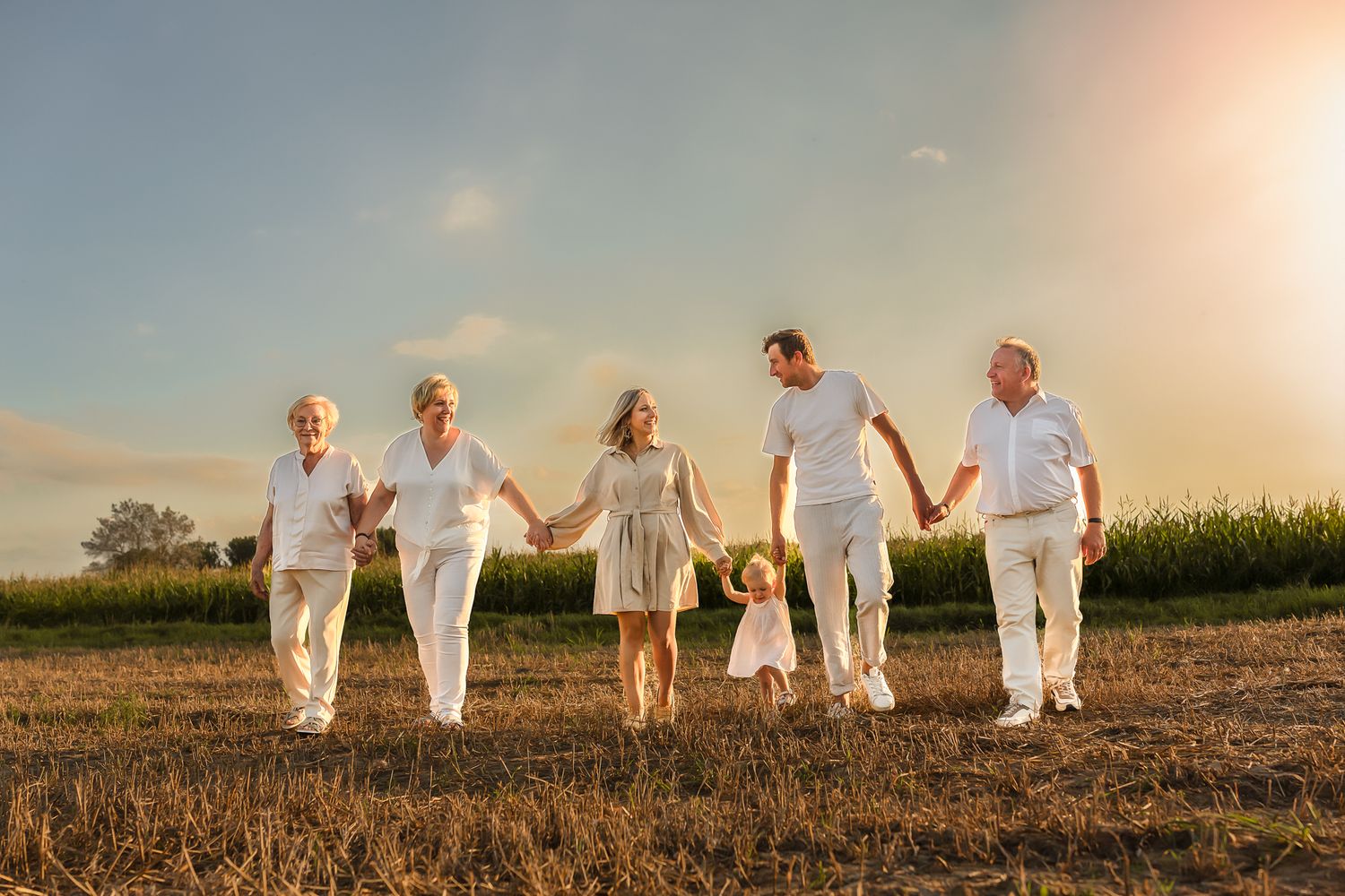 Multi-generational family dressed in white holds hands while walking through rural field at sunset.