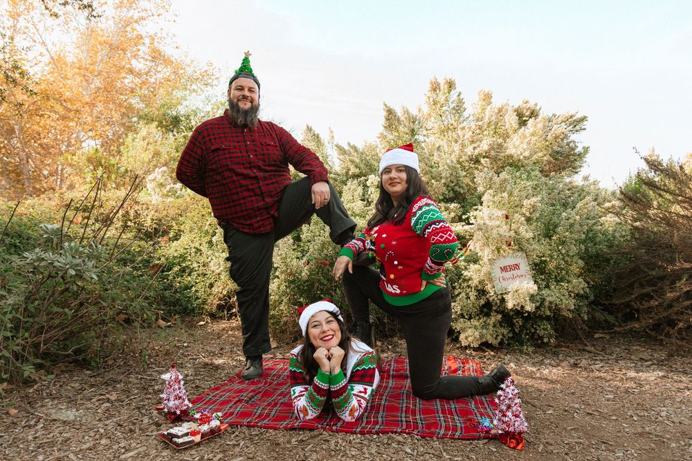 Playful holiday photoshoot outdoors with matching Christmas sweaters and Santa hats on red blanket.