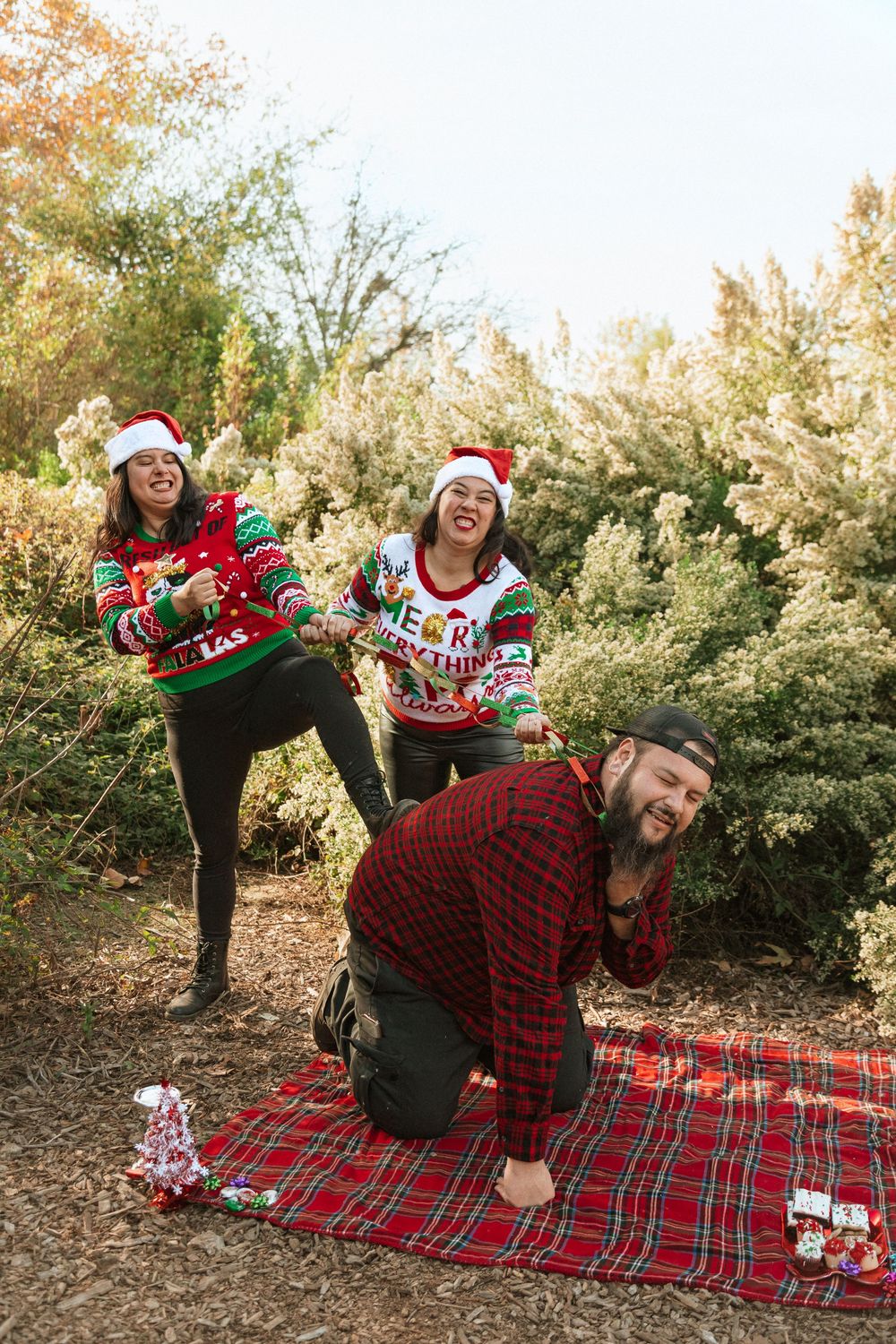 Three people strike funny poses on a red plaid blanket while wearing festive Christmas sweaters and Santa hats.