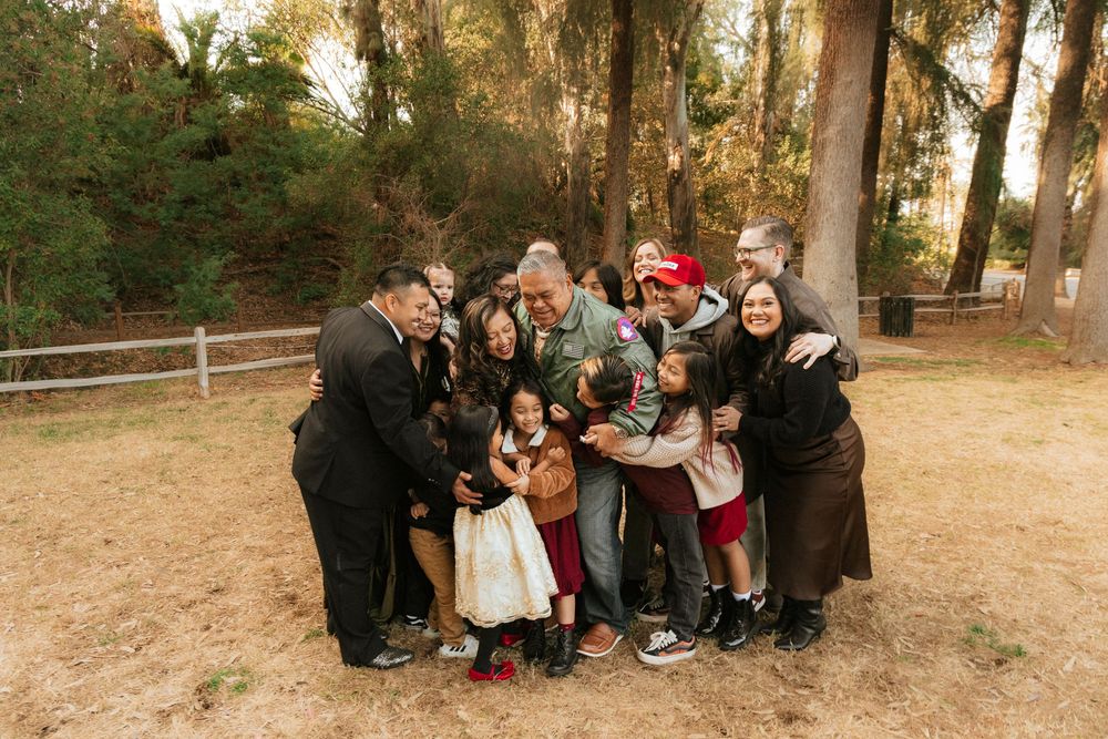 Large group gathering in a sun-dappled forest clearing with people arranged casually near tall trees.