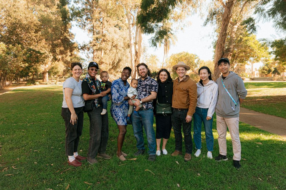 A group of diverse friends stand together smiling on a grassy lawn with tall trees in the background on a sunny day.