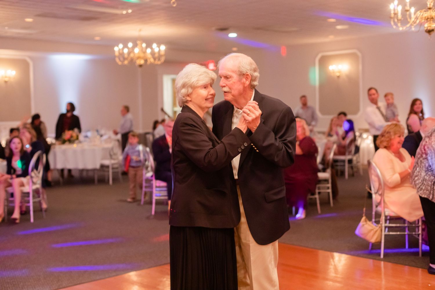Ballroom dance party with couples on the dance floor under crystal chandeliers.