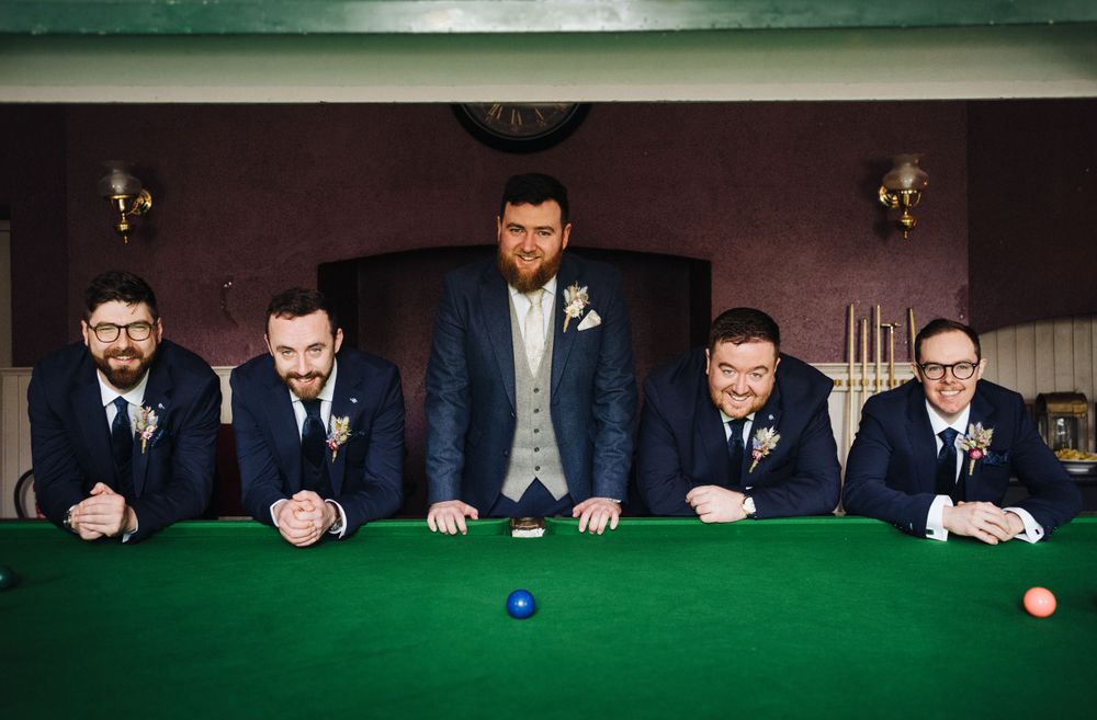 Groomsmen gathered around green felt pool table for formal wedding party photo.