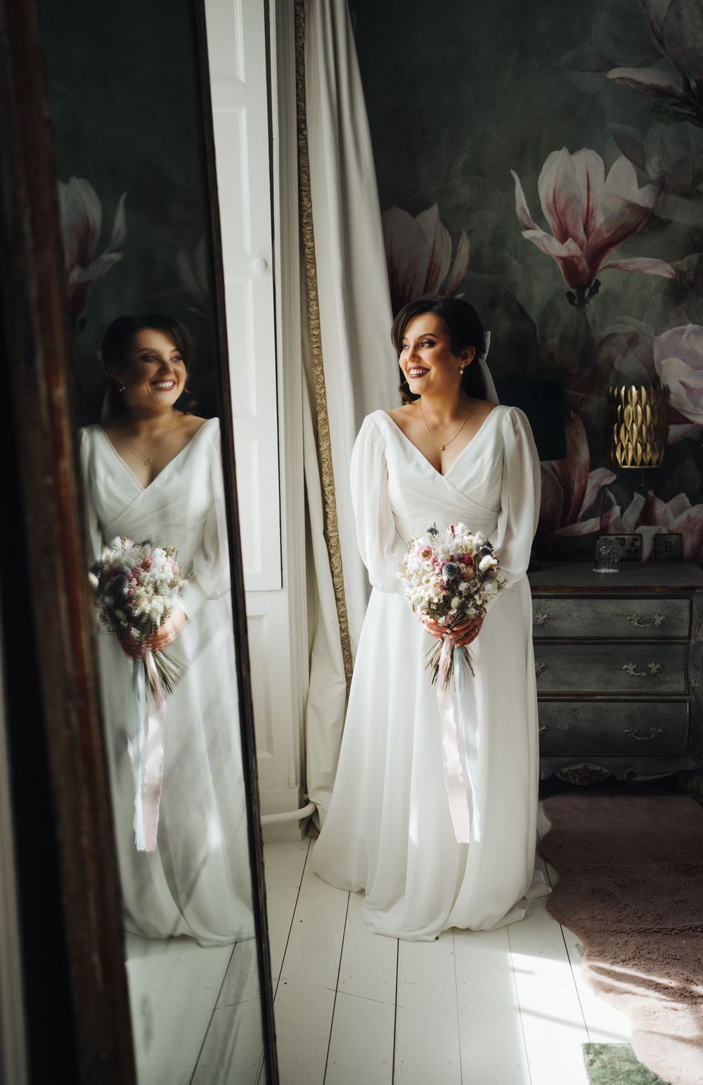 Bride in elegant long-sleeve wedding dress poses beside mirror with floral wallpaper background.