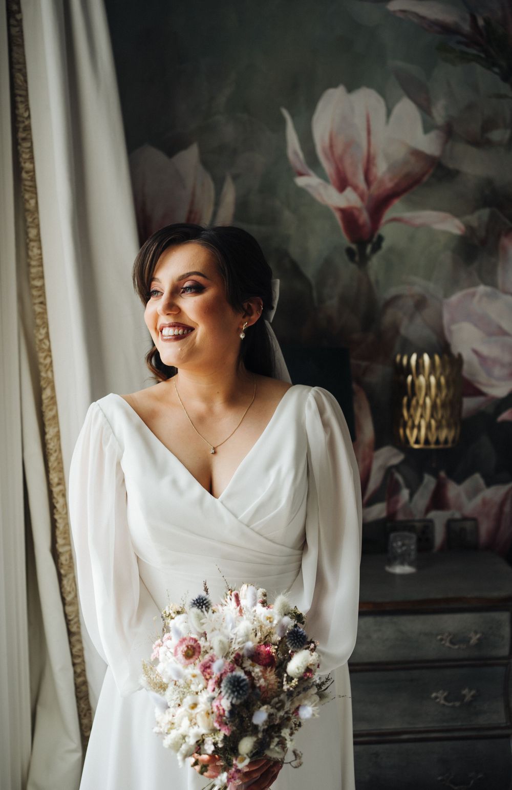 Bride holding pastel bouquet poses by window with magnolia flower wallpaper backdrop.