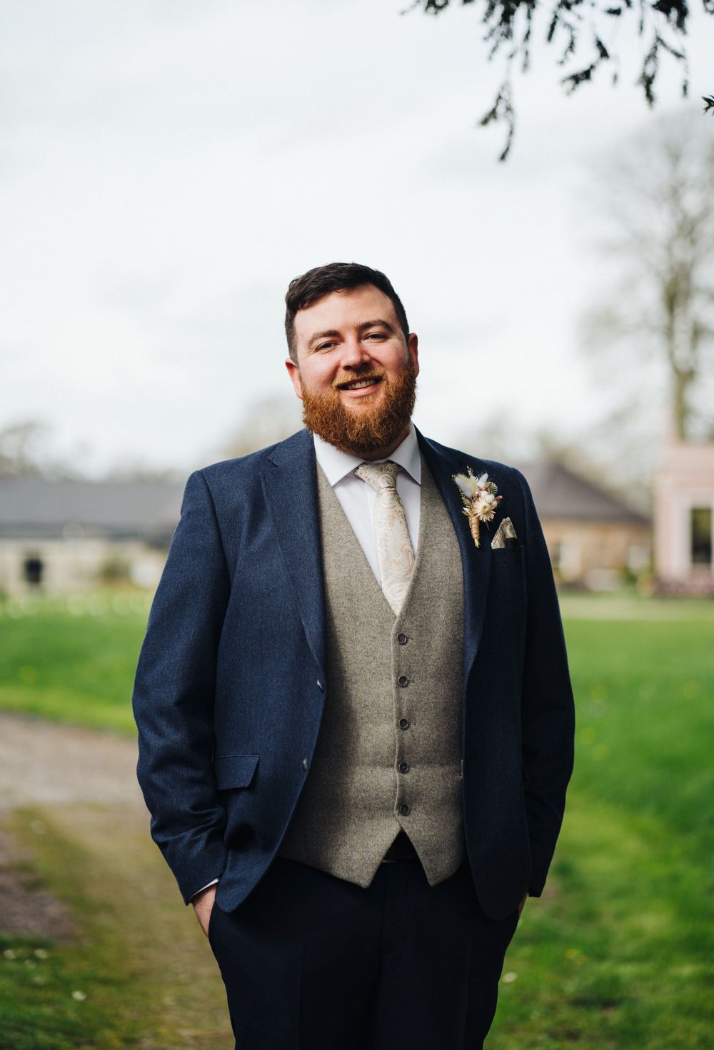 Groom in navy suit and grey waistcoat poses outdoors on wedding day.
