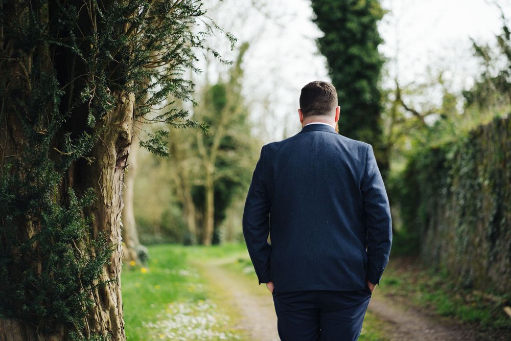 Back view of person in navy suit walking along garden path surrounded by greenery.