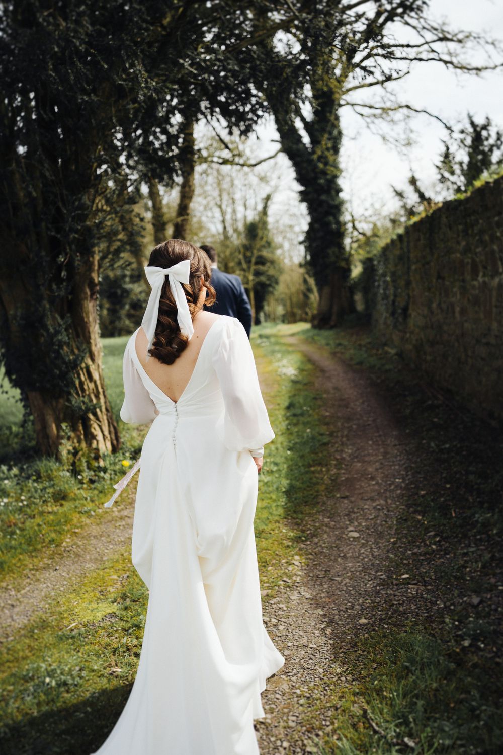 Bride in flowing white dress walking along tree-lined countryside path.