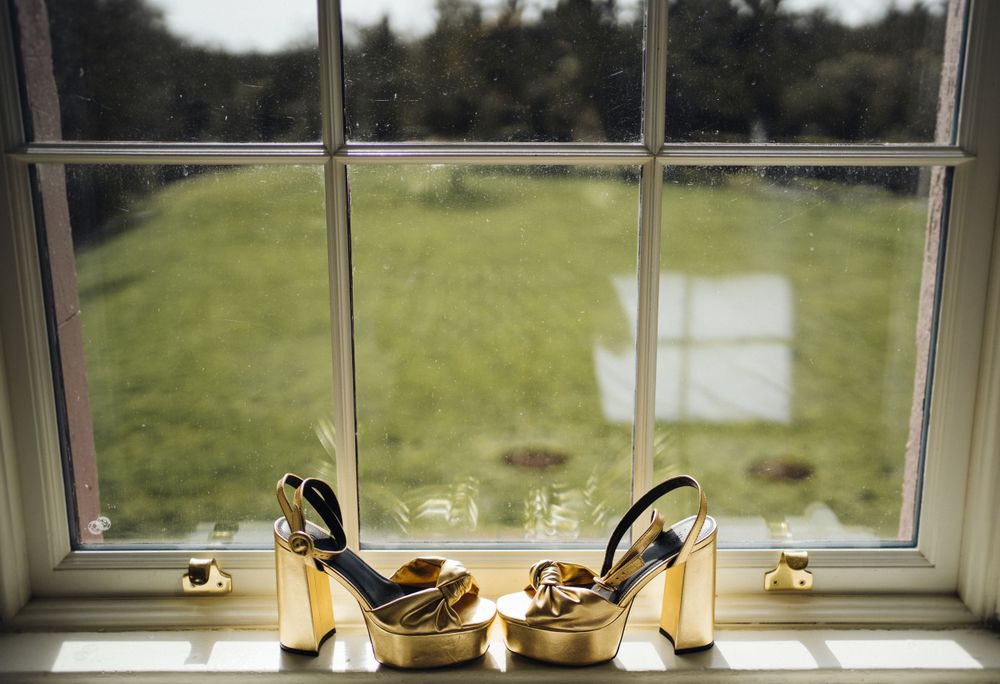 Gold wedding shoes displayed on a windowsill overlooking a green lawn.