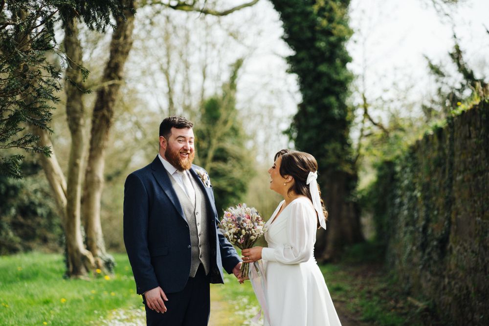 A couple on their wedding day stands together holding a floral bouquet on a green path.