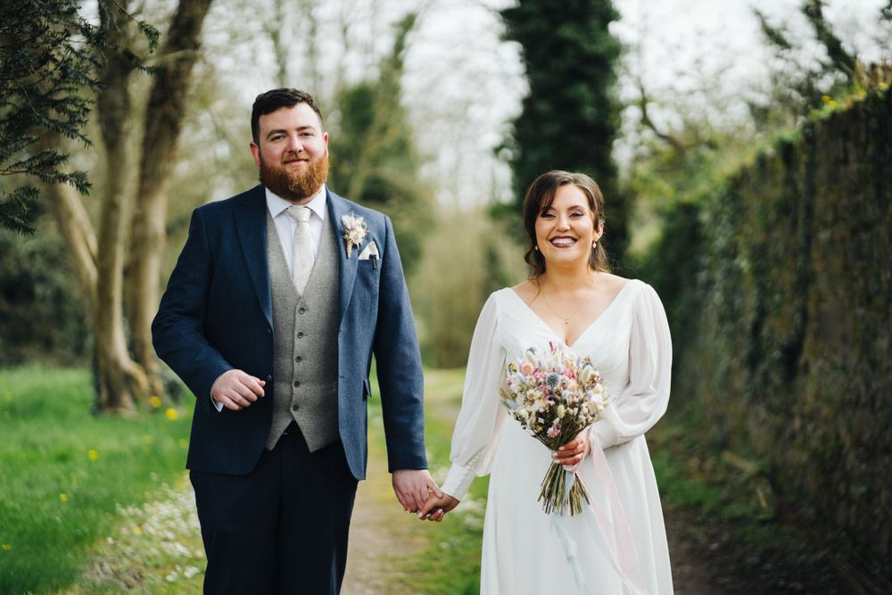 A joyful newlywed couple shares a laugh while holding hands on a garden walkway.