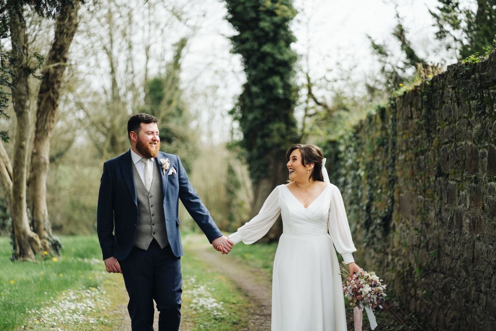 Newlyweds walk together on a stone-walled garden path during their spring wedding.