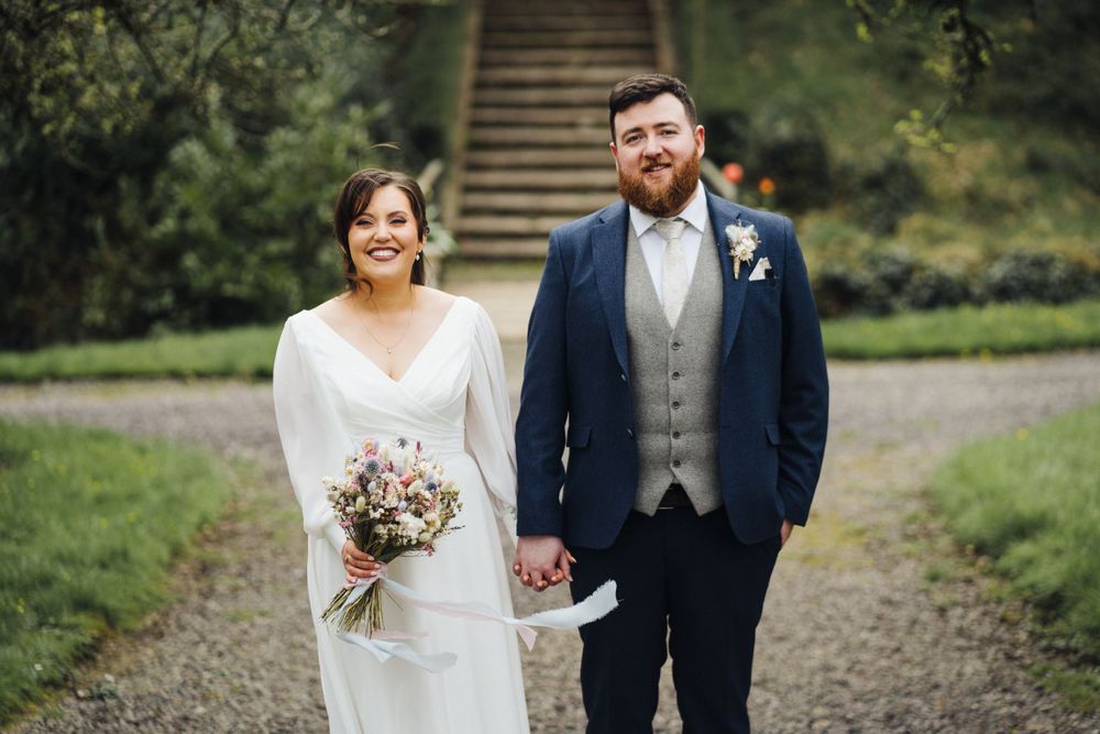 Newlywed couple walking together on gravel path with bride holding romantic bouquet.