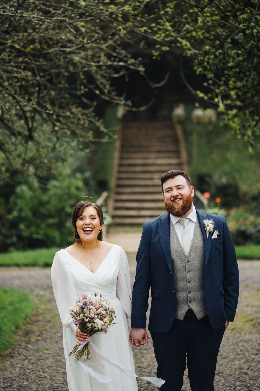 A joyful wedding couple stands on a stone path in front of rustic stairs surrounded by lush greenery.