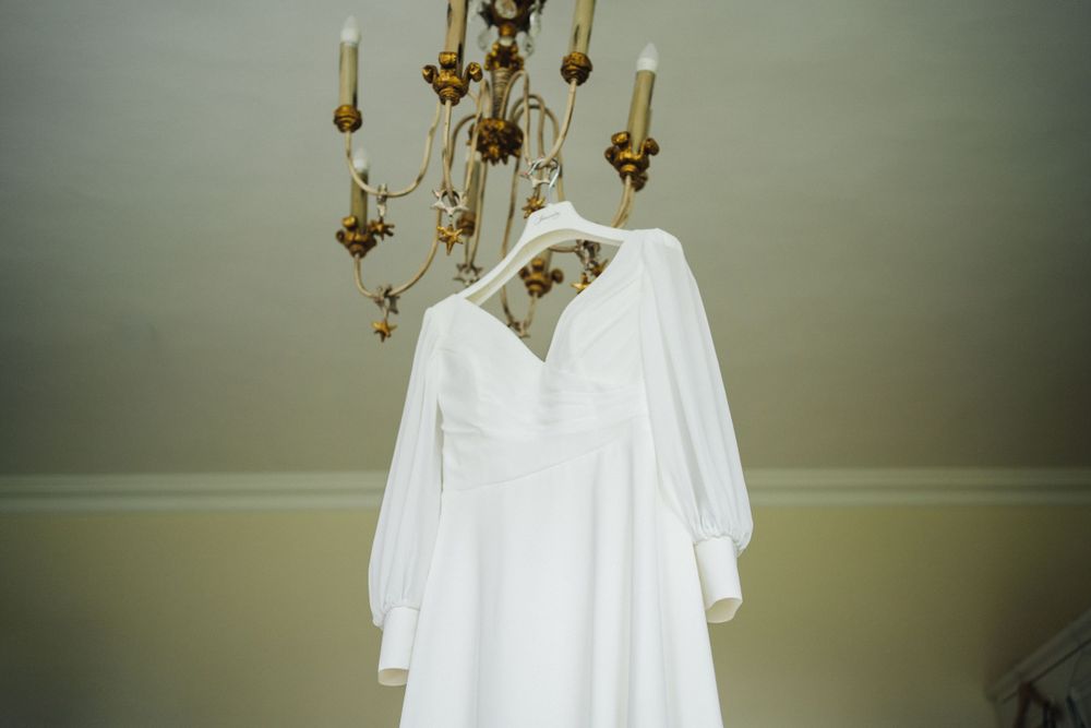 A white wedding dress hanging from an elegant chandelier in a neutral-toned room.