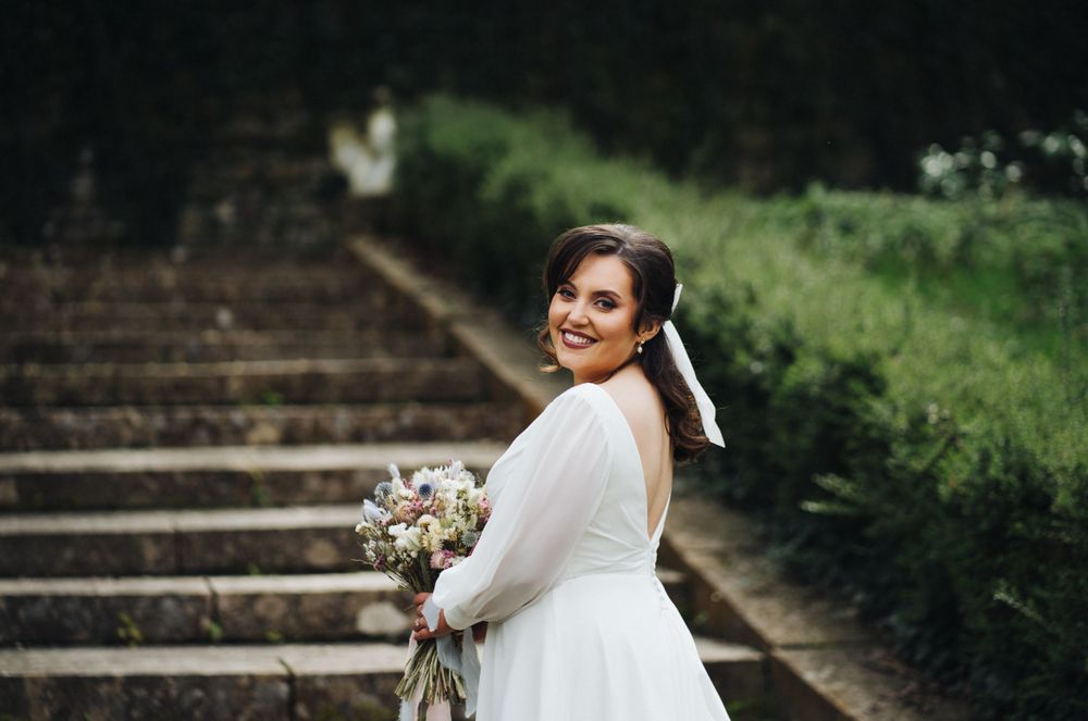 A bride in a white dress poses on stone garden steps while holding a pastel bouquet.