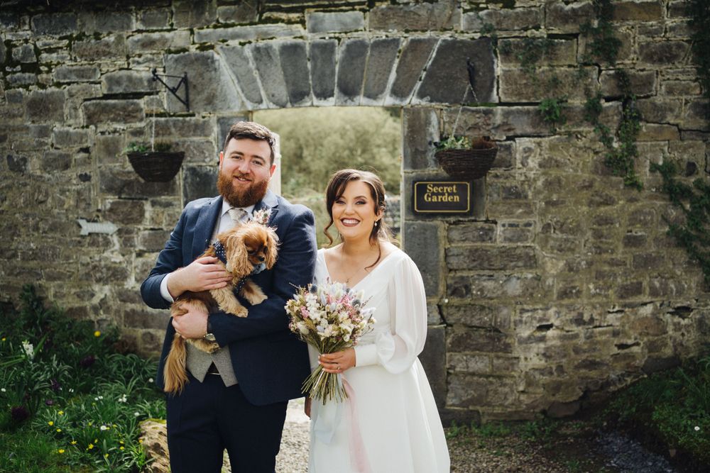 A couple poses with their small dog by a stone archway during their wedding.