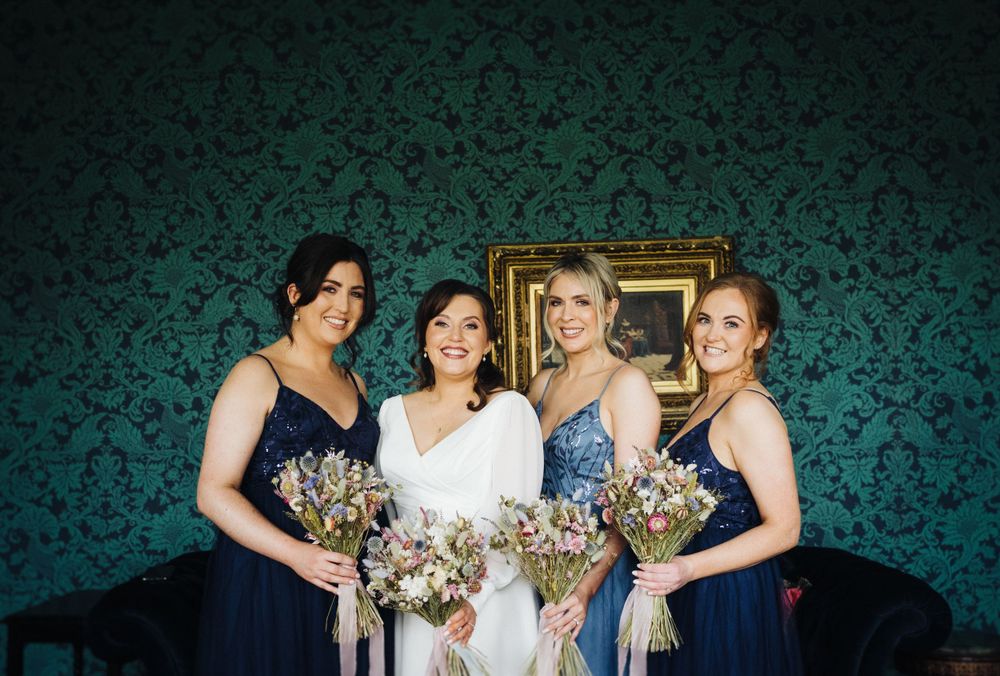 Bridesmaids in blue dresses pose against a teal damask wall holding bouquets.