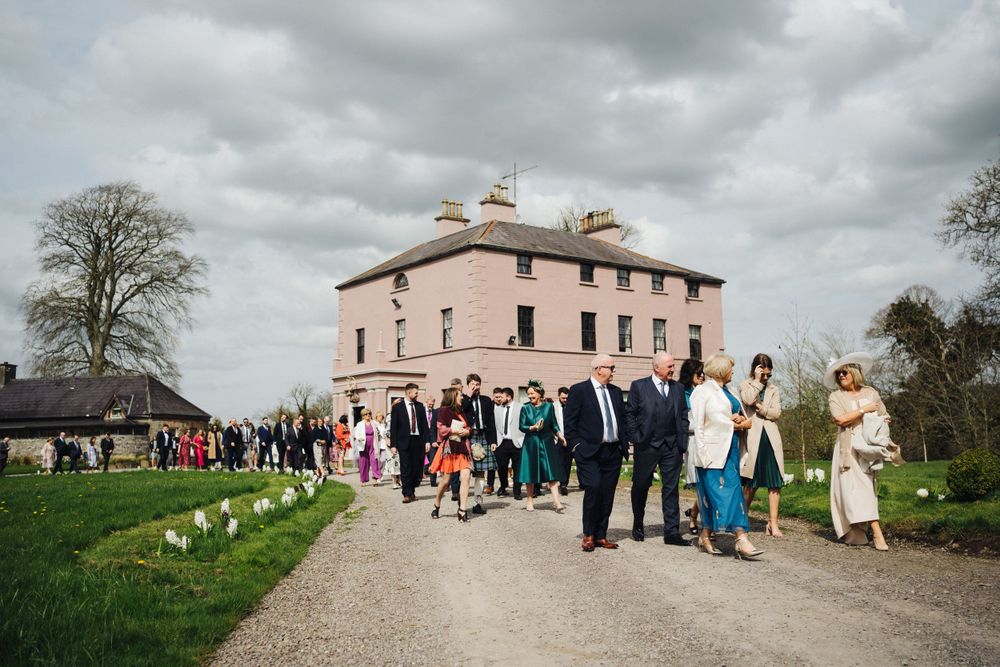 Wedding guests gather outside a pink historic mansion on a cloudy day.