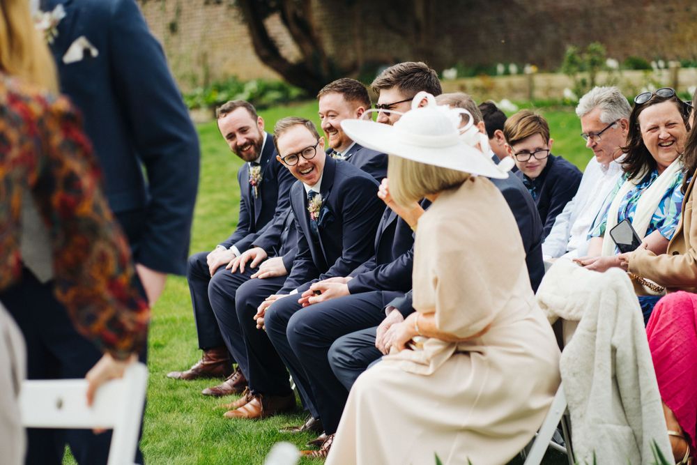 Wedding ceremony guests laugh while seated in rows on a green lawn.