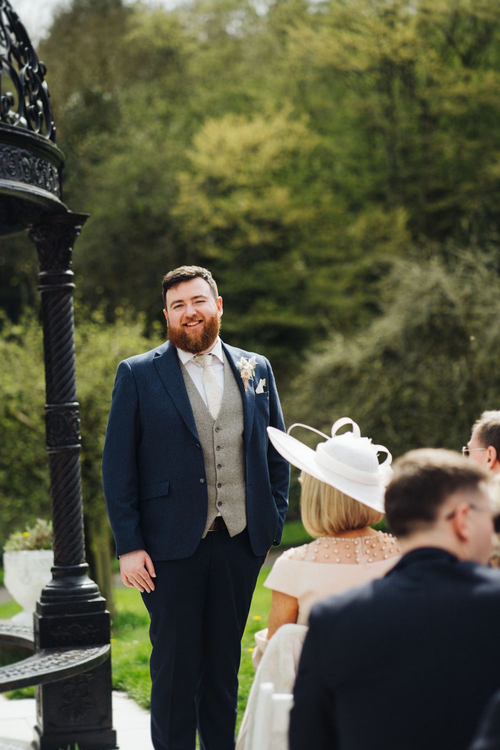 Elegantly dressed groomsman in navy suit and bowtie stands near decorative lamp post at outdoor wedding ceremony.