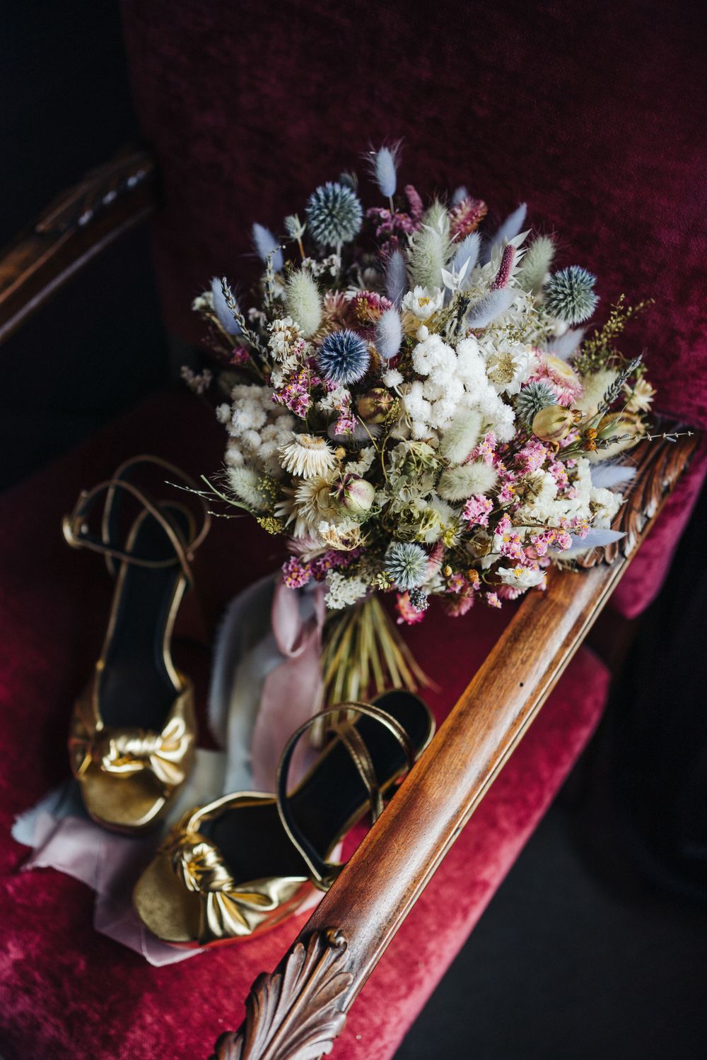 A vintage-style dried flower wedding bouquet with gold shoes on red velvet furniture.