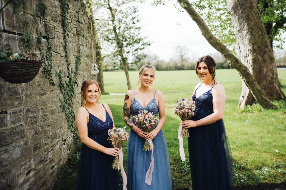 Three bridesmaids in navy and light blue dresses pose with bouquets against stone wall outdoors.