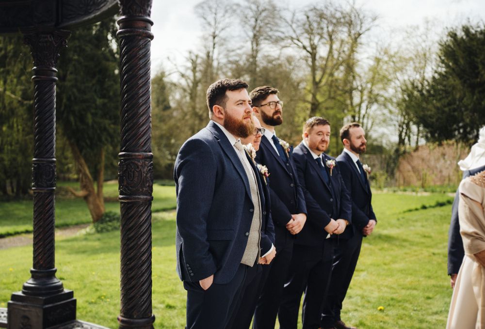 Groomsmen lined up in matching navy suits during outdoor garden wedding ceremony.
