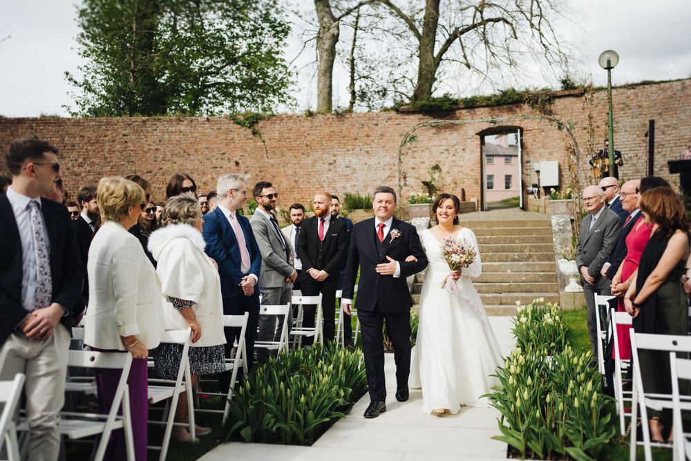Wedding ceremony processional on stone steps with brick wall backdrop and white garden chairs.