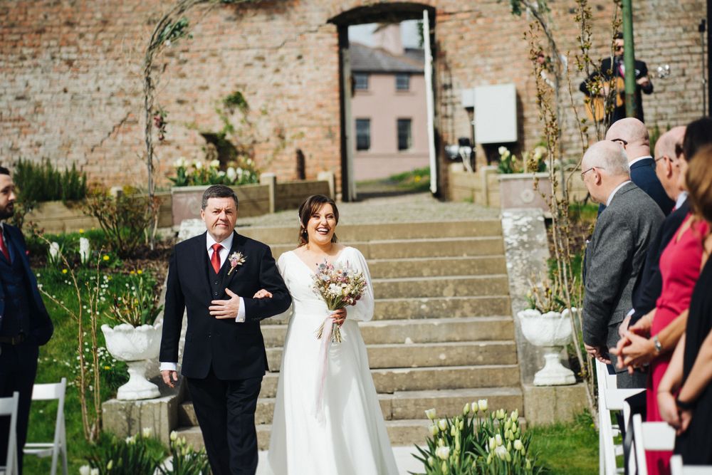 Couple walks down outdoor stone staircase during wedding ceremony with guests seated on white chairs.