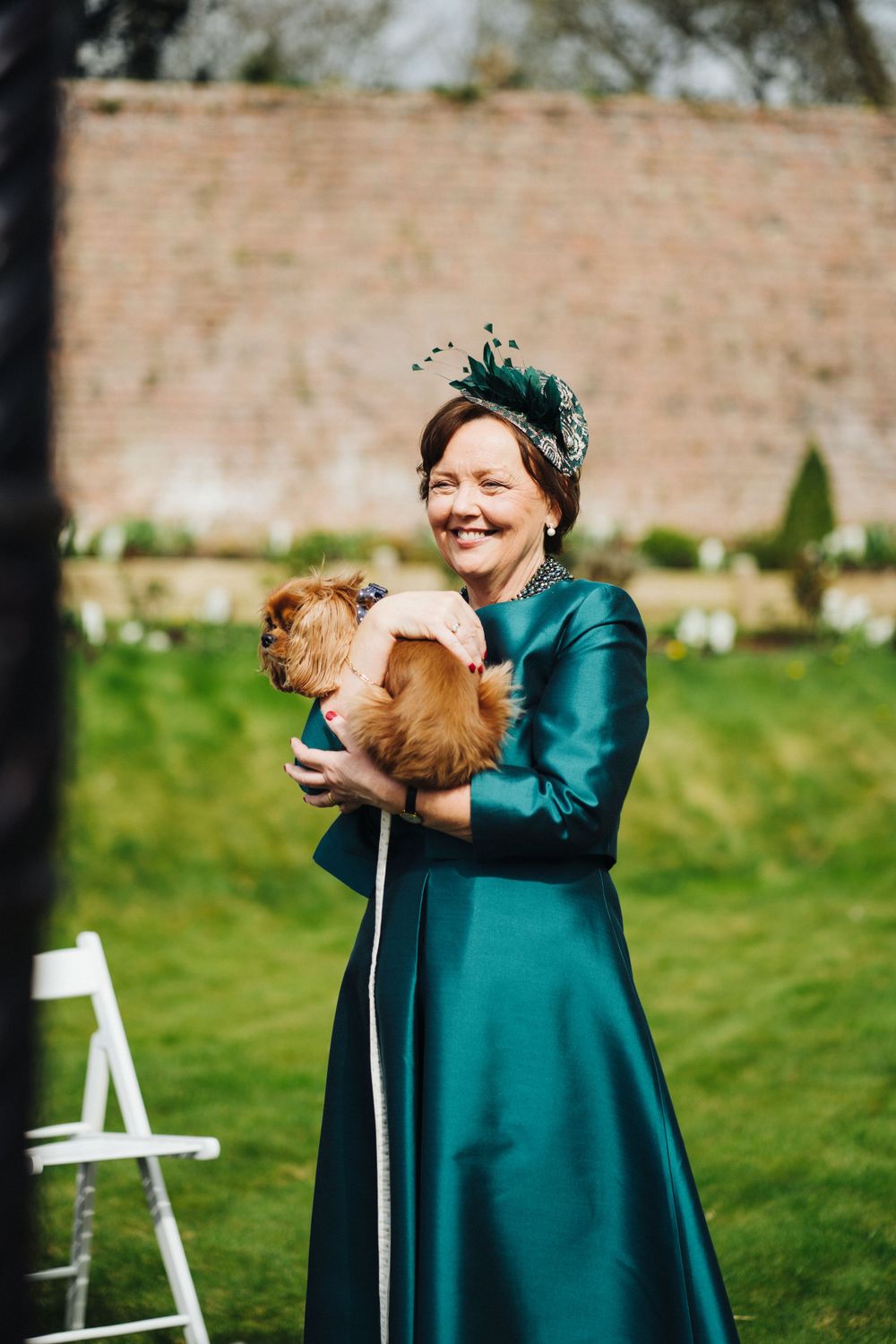 A small dog being held at an outdoor wedding event against a brick wall backdrop.