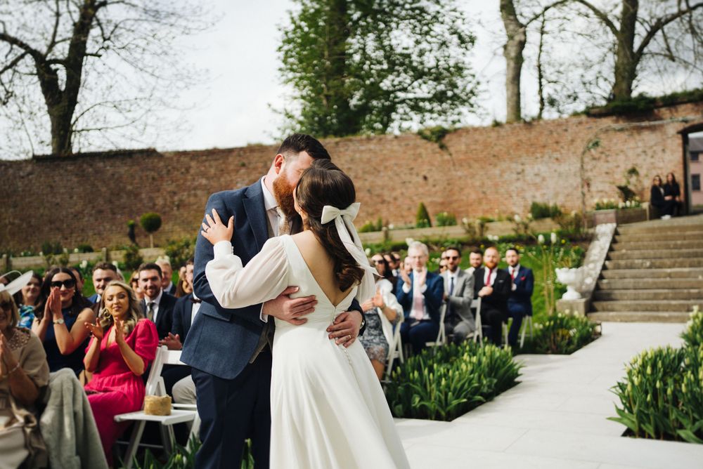 A romantic wedding ceremony kiss in front of gathered guests at an outdoor garden venue.